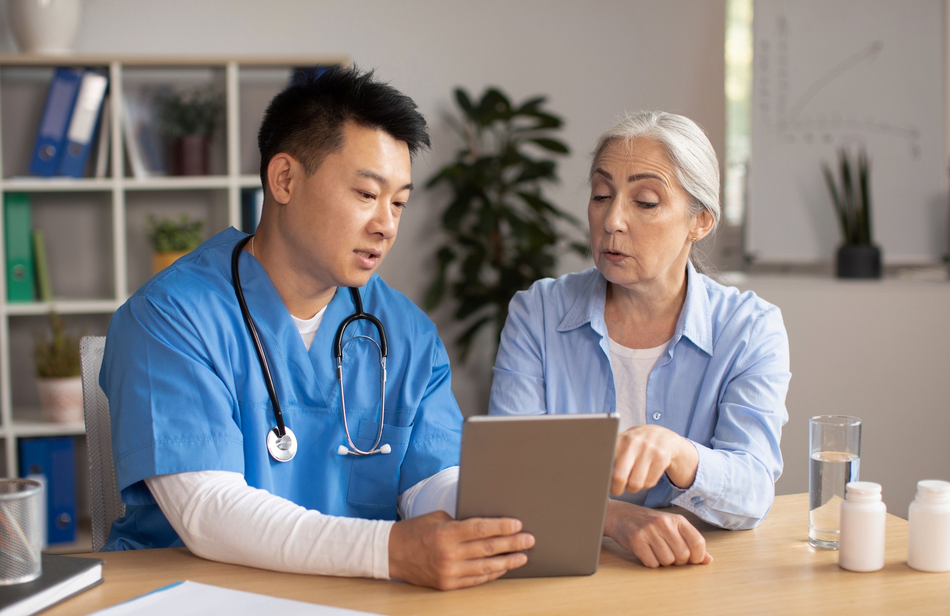 Serious middle aged korean male doctor showing tablet and consulting senior european lady patient in clinic